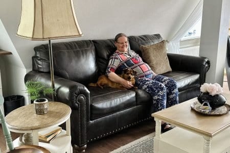 An elderly man wearing glasses and a light shirt is reclining with eyes closed in a sunlit room. A woman sits nearby looking ahead. A couch and a vase with flowers are in the background. The ambiance is peaceful and relaxed.