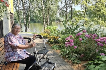 People in a park performing seated exercises near a pond. One woman stands with arms raised, guiding others. They are under patio umbrellas. The setting is lush and green with clear skies and bright sunlight.