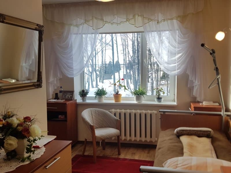 A cozy bedroom with a hospital bed, wooden furniture, and a large window adorned with sheer curtains. Potted plants sit on the windowsill, and there's a scenic view of trees and snow outside. A flower arrangement is on the dresser.