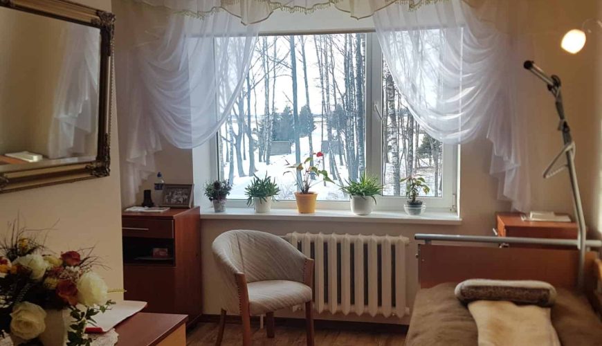 A cozy bedroom with a hospital bed, wooden furniture, and a large window adorned with sheer curtains. Potted plants sit on the windowsill, and there's a scenic view of trees and snow outside. A flower arrangement is on the dresser.