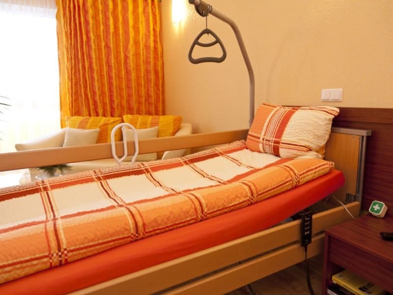 A cozy bedroom with an adjustable bed featuring an orange and white checkered blanket and pillow. A beige wall with orange curtains and a small sitting area with cream and yellow cushions in the background. A green first aid kit sits on a side table.