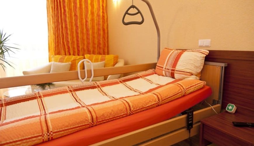 A cozy bedroom with an adjustable bed featuring an orange and white checkered blanket and pillow. A beige wall with orange curtains and a small sitting area with cream and yellow cushions in the background. A green first aid kit sits on a side table.