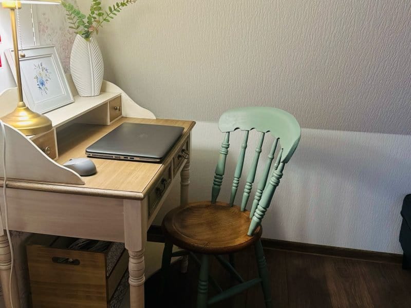 A cozy workspace featuring a vintage wooden desk with a laptop, a mouse, and a potted plant. A green wooden chair is placed at the desk. A table lamp emits a warm glow, illuminating a framed floral artwork on the wall.