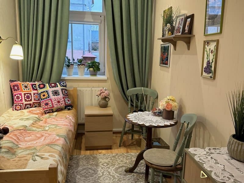 Cozy room with a single bed featuring colorful crochet pillows and a floral blanket. A small round table with chairs sits in the corner. Green curtains frame the window, and shelves with plants and photos adorn the walls. A chest of drawers is nearby.