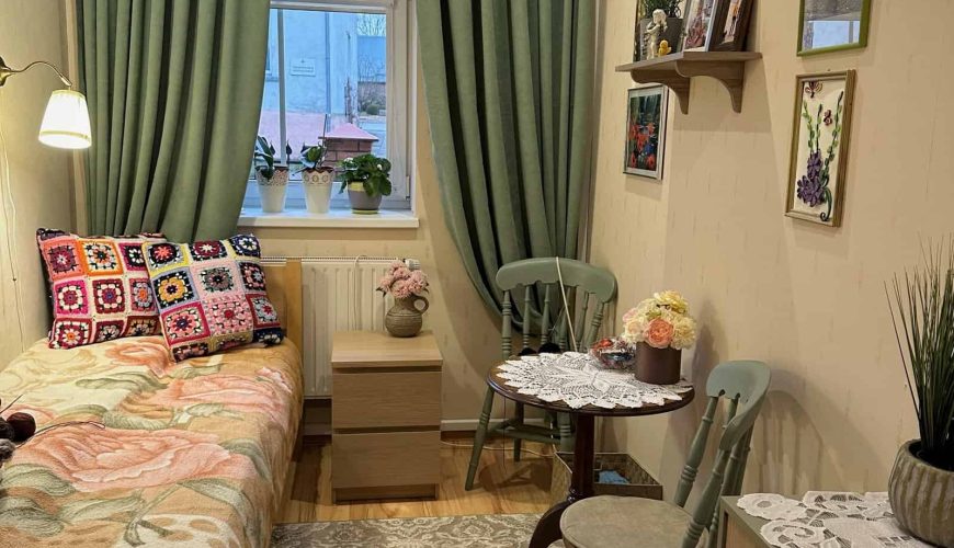 Cozy room with a single bed featuring colorful crochet pillows and a floral blanket. A small round table with chairs sits in the corner. Green curtains frame the window, and shelves with plants and photos adorn the walls. A chest of drawers is nearby.