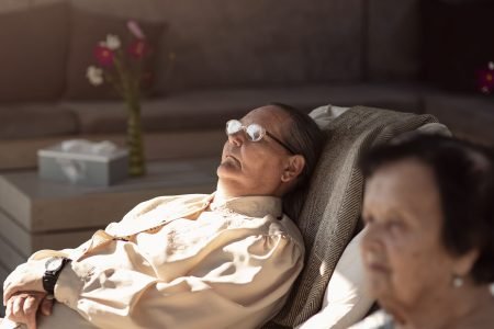 An elderly man wearing glasses and a light shirt is reclining with eyes closed in a sunlit room. A woman sits nearby looking ahead. A couch and a vase with flowers are in the background. The ambiance is peaceful and relaxed.