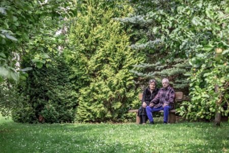 An elderly couple sits together on a wooden bench in a lush, green garden. They are surrounded by tall trees and foliage, and appear to be smiling. The grass is vibrant, and the scene is peaceful and natural.