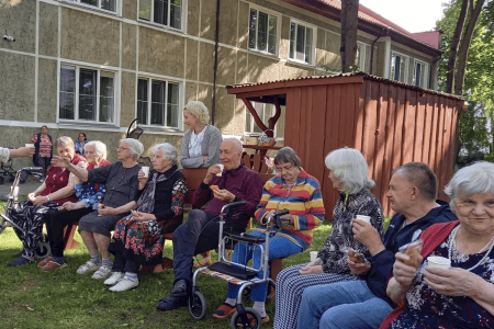 A group of elderly people sitting on benches and in wheelchairs outside, enjoying ice cream. They are in a grassy area with trees and a building in the background. It's a sunny day and everyone appears to be relaxed and smiling.
