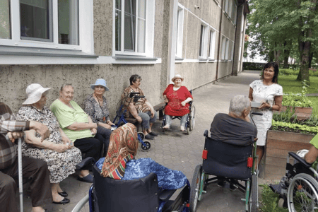 A group of elderly people sitting on benches and in wheelchairs outside, enjoying ice cream. They are in a grassy area with trees and a building in the background. It's a sunny day and everyone appears to be relaxed and smiling.