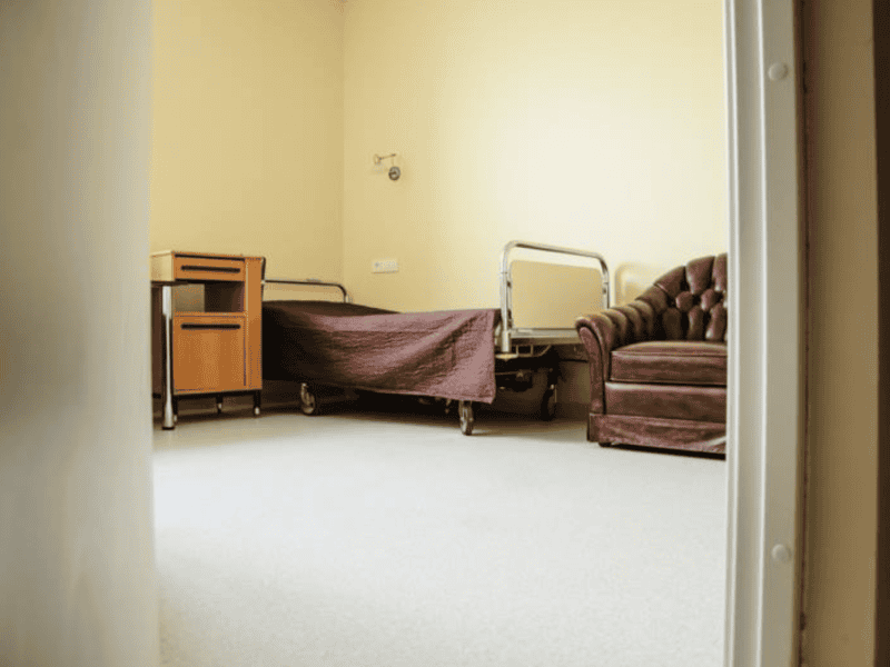 A simple hospital room with a bed covered in a brown blanket, a wooden bedside table, and a brown leather armchair. The room has beige walls and a light gray floor, viewed from an open doorway.