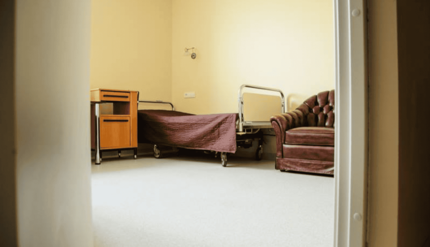 A simple hospital room with a bed covered in a brown blanket, a wooden bedside table, and a brown leather armchair. The room has beige walls and a light gray floor, viewed from an open doorway.