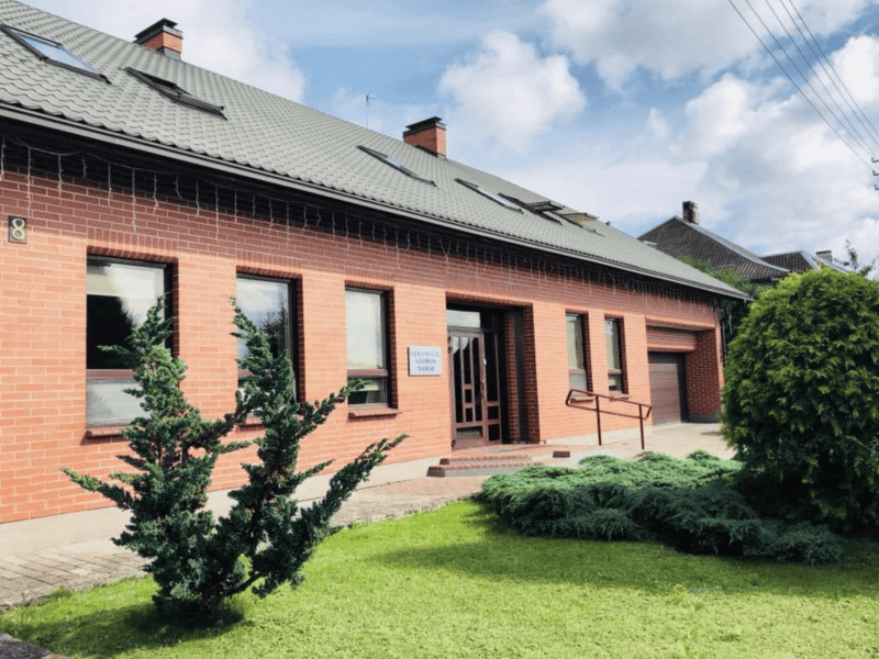 A single-story red-brick building with a sloped gray roof and several windows. It has a wooden door, a small sign beside it, and a wheelchair ramp. A well-maintained lawn with shrubs is in the foreground under a partly cloudy sky.