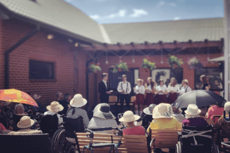 A group of elderly people sits around a long outdoor table, enjoying beverages. Colorful umbrellas hang overhead as decoration. The setting is a sunlit patio with brick walls and part of a building visible in the background.