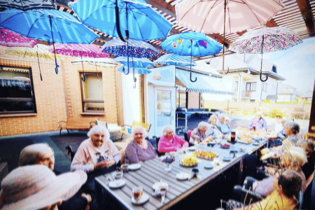 A group of elderly people sits around a long outdoor table, enjoying beverages. Colorful umbrellas hang overhead as decoration. The setting is a sunlit patio with brick walls and part of a building visible in the background.
