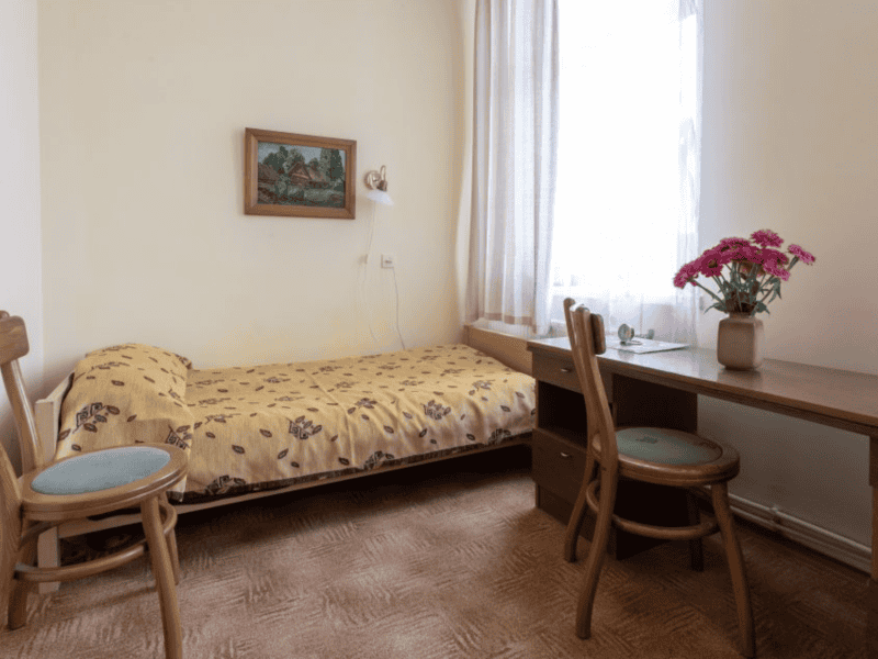 A small, cozy room with a single bed covered in a floral blanket, a wooden desk with a vase of pink flowers, and two chairs. A painting hangs above the bed, and light filters through a window with sheer curtains.