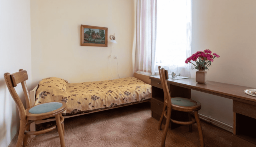 A small, cozy room with a single bed covered in a floral blanket, a wooden desk with a vase of pink flowers, and two chairs. A painting hangs above the bed, and light filters through a window with sheer curtains.