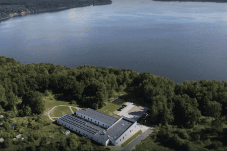 Aerial view of a large, rectangular building with solar panels on the roof, surrounded by dense green forest. The building is near the shore of a calm, expansive lake, with a parking area and a few vehicles visible. The horizon shows a tree-lined coastline.