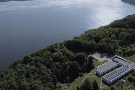 Aerial view of a large, rectangular building with solar panels on the roof, surrounded by dense green forest. The building is near the shore of a calm, expansive lake, with a parking area and a few vehicles visible. The horizon shows a tree-lined coastline.