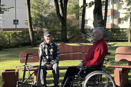 A group of elderly people sitting on benches and in wheelchairs outside, enjoying ice cream. They are in a grassy area with trees and a building in the background. It's a sunny day and everyone appears to be relaxed and smiling.