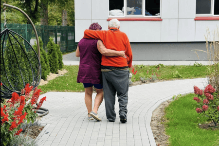 A group of elderly people sitting on benches and in wheelchairs outside, enjoying ice cream. They are in a grassy area with trees and a building in the background. It's a sunny day and everyone appears to be relaxed and smiling.