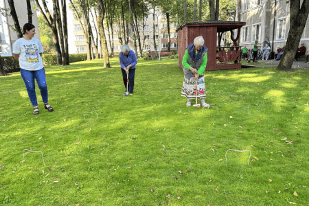 A group of elderly people sitting on benches and in wheelchairs outside, enjoying ice cream. They are in a grassy area with trees and a building in the background. It's a sunny day and everyone appears to be relaxed and smiling.