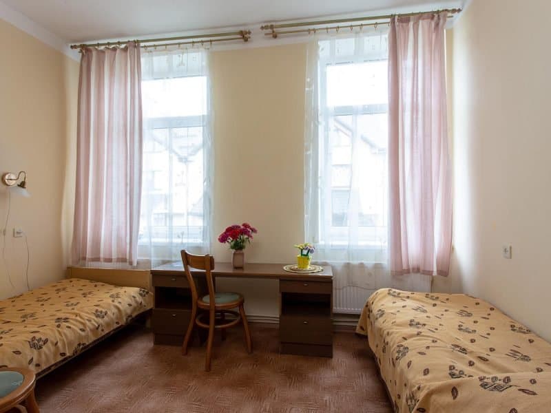 A small room with two single beds on either side, separated by a wooden desk and chair. Pink curtains cover two tall windows. A vase with red flowers and a cup with yellow flowers sit on the desk. The bedspreads have a floral pattern.