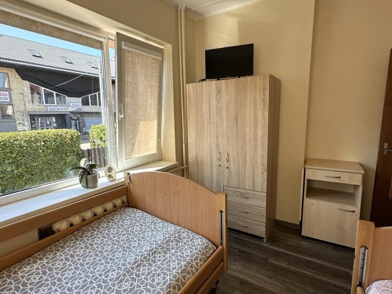 A small room featuring two wooden single beds with patterned mattresses. A window lets in natural light, illuminating a wooden wardrobe with a TV on top. A small nightstand is beside the wardrobe. The view includes a hedge and buildings outside.