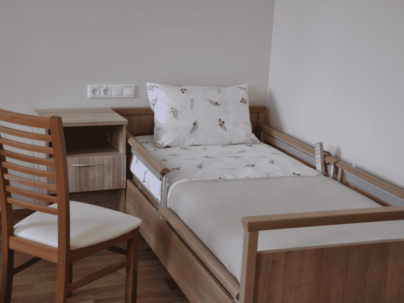 A neatly made hospital bed with white floral sheets and a pillow is adjacent to a wooden bedside table. A wooden chair with a cream cushion is positioned next to the table. The room has a clean, simple design.