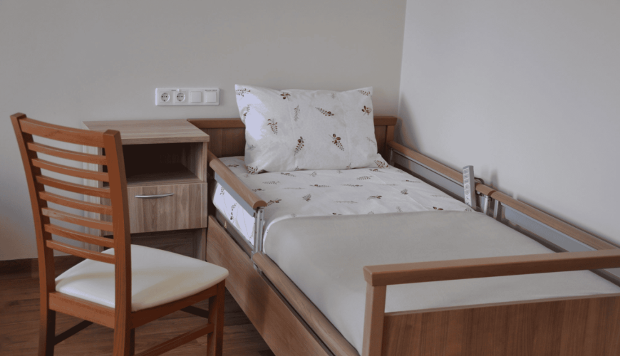 A neatly made hospital bed with white floral sheets and a pillow is adjacent to a wooden bedside table. A wooden chair with a cream cushion is positioned next to the table. The room has a clean, simple design.