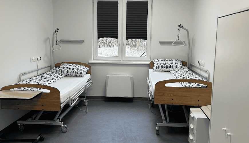 A hospital room with two empty beds featuring wood paneling, patterned bedding, overbed tables, and wall-mounted reading lamps. The room has a window with blinds, white walls, and a gray floor. A cabinet and a heater are also visible.