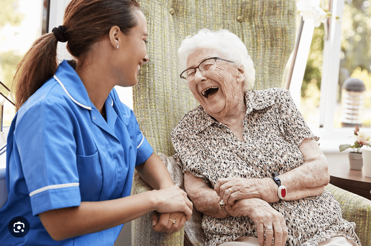 A nurse in a blue uniform shares a joyful moment with an elderly woman wearing glasses and a patterned blouse. They are seated in a bright room, smiling and laughing together.