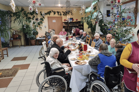 A group of elderly people, some seated and some in wheelchairs, pose outside a building under a bright blue sky. Many are holding balloons and wearing hats, with a well-kept garden and part of a patio visible in the background.