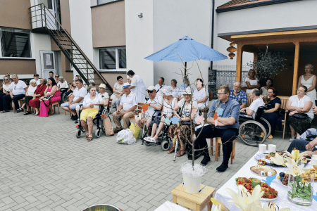 A group of elderly people, some seated and some in wheelchairs, pose outside a building under a bright blue sky. Many are holding balloons and wearing hats, with a well-kept garden and part of a patio visible in the background.