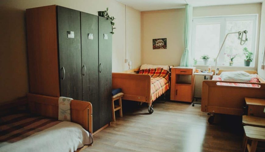 A cozy room with three wooden beds, a large dark wardrobe, light walls, a window with green curtains, potted plants, a small table, and soft natural light filling the space.