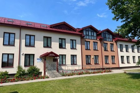 A two-story building with red roofs and cream and maroon walls stands under a blue sky. It has multiple windows and a central entrance with a ramp. The front yard features a path, a green lawn, and red flowers.