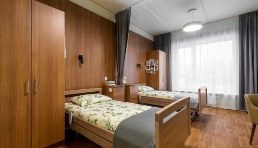 A bright, tidy room with two single beds featuring floral bedding, wooden headboards, matching wardrobes, a desk, a chair, and large windows with sheer curtains letting in natural light.