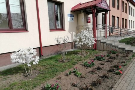 A two-story building with red roofs and cream and maroon walls stands under a blue sky. It has multiple windows and a central entrance with a ramp. The front yard features a path, a green lawn, and red flowers.