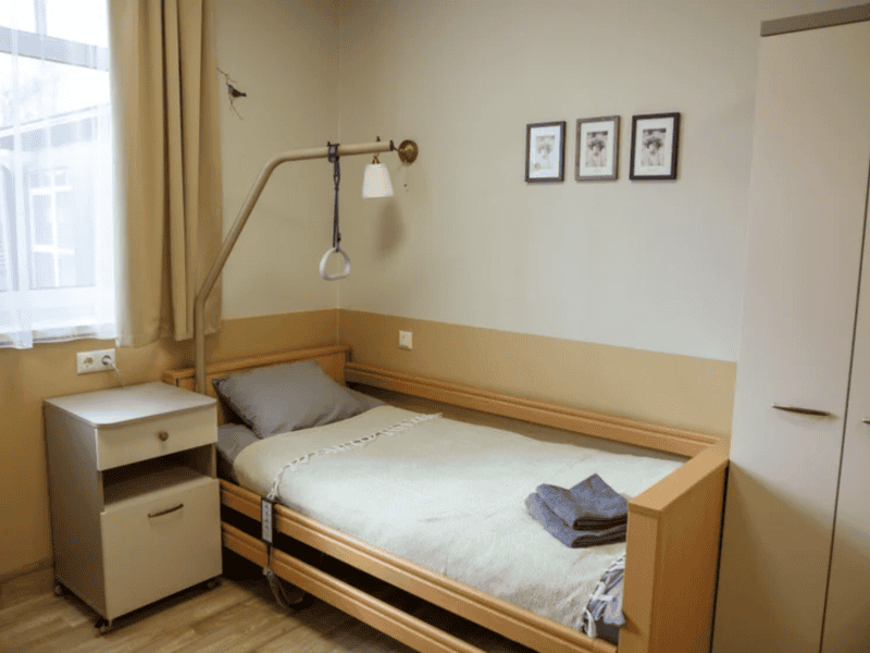 A neatly arranged single hospital room with a bed featuring a green blanket and two towels. A bedside table, a wall-mounted light with a handle, a window with curtains, and three framed pictures on the wall are visible.