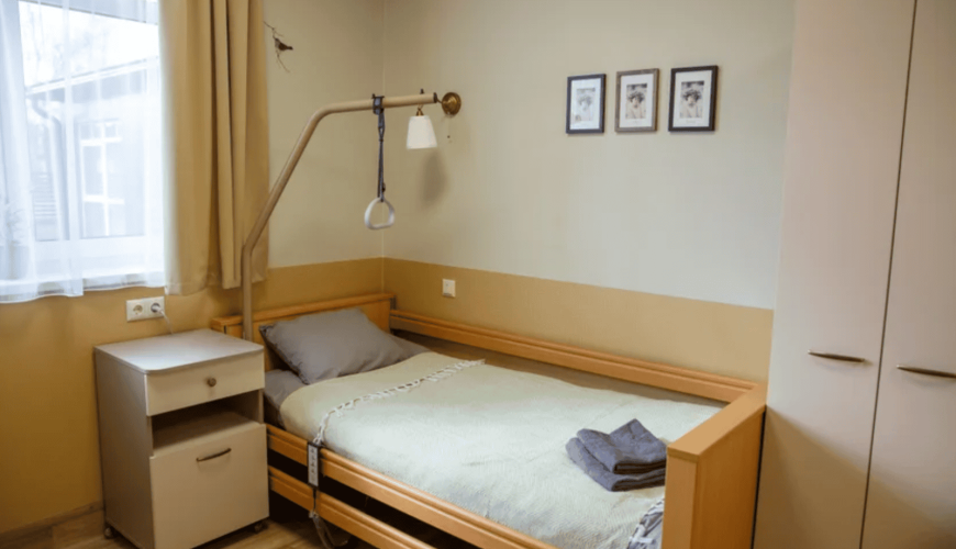 A neatly arranged single hospital room with a bed featuring a green blanket and two towels. A bedside table, a wall-mounted light with a handle, a window with curtains, and three framed pictures on the wall are visible.