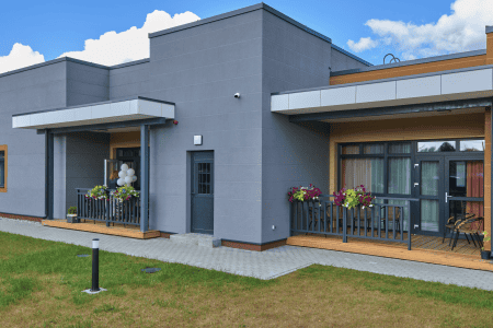 Modern single-story building with gray exterior and flat roof. Two covered porches display flower boxes and balloons. The setting is grassy with a paved path. The sky is partly cloudy, adding contrast to the sleek architecture.