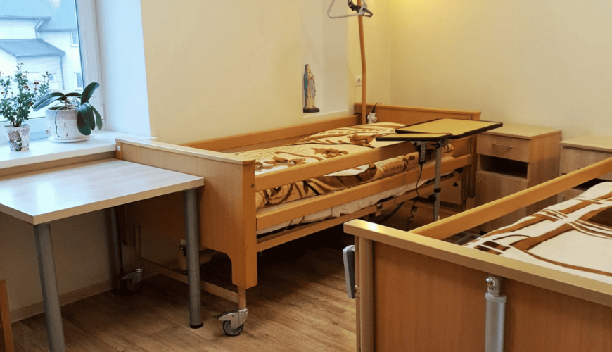 A tidy room with two adjustable hospital beds and wooden furniture. There's a window with potted plants on a table, and a lamp mounted on the wall. The flooring is laminate, and there's a serene atmosphere.