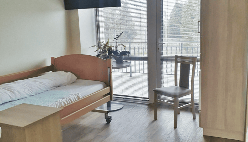 A clean, simple room with a single bed, wooden chair, small table, and a TV mounted on the wall. A large window with blinds overlooks a balcony. A plant sits on the window ledge, adding a touch of greenery to the space.