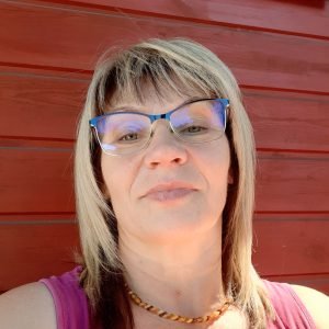 A woman with blonde hair and glasses wears a pink sleeveless top and a beaded necklace, standing in front of a red wooden wall, looking at the camera and smiling slightly.