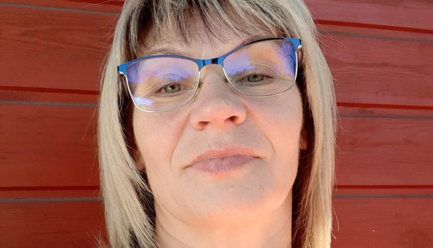 A woman with blonde hair and glasses wears a pink sleeveless top and a beaded necklace, standing in front of a red wooden wall, looking at the camera and smiling slightly.