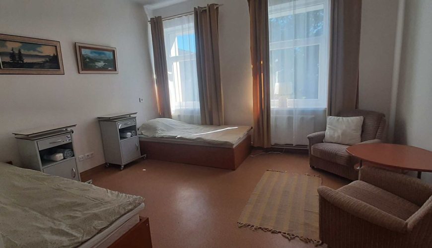 A simple room with two single beds, two nightstands, two armchairs, a small round table, a rug, and two large windows with curtains; landscape paintings hang on the white walls.