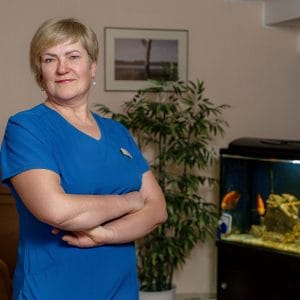 A woman in blue scrubs stands with arms crossed, smiling confidently in a room with a fish tank, a potted plant, and a framed picture on the wall.