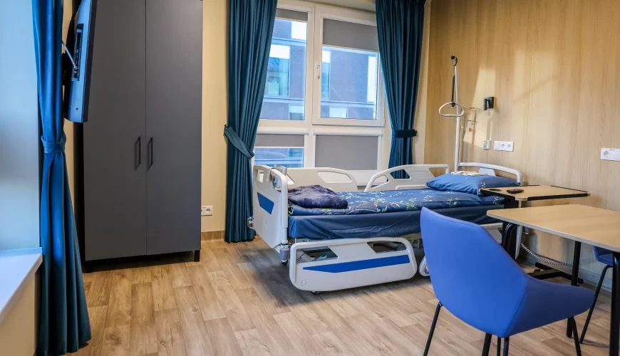 A modern hospital room with a single adjustable bed, blue bedding, a table and chair, a wardrobe, a wall-mounted TV, and large windows with blue curtains. The floor is light wood, and the walls are light-colored.