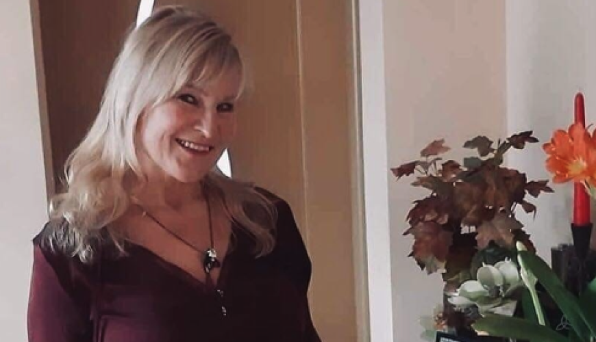 A smiling woman with long blonde hair wearing a burgundy top and plaid pants stands indoors beside a wooden sideboard decorated with potted plants and a framed photo.