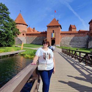 Woman wearing sunglasses and a white shirt stands on a wooden bridge in front of a large red-brick castle with towers, under a clear blue sky. Other visitors and greenery are visible in the background.