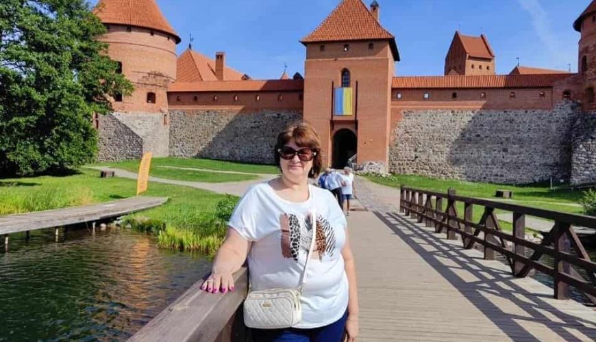 Woman wearing sunglasses and a white shirt stands on a wooden bridge in front of a large red-brick castle with towers, under a clear blue sky. Other visitors and greenery are visible in the background.